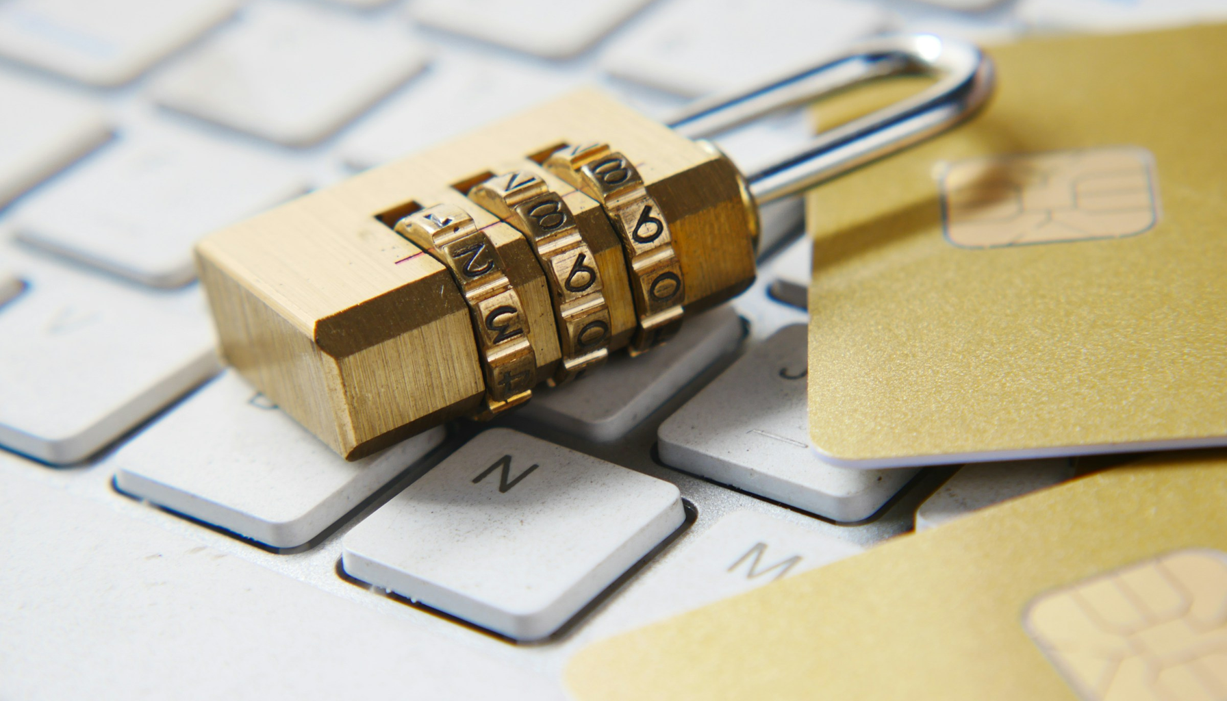 A gold combination padlock resting on a white computer keyboard next to gold payment cards