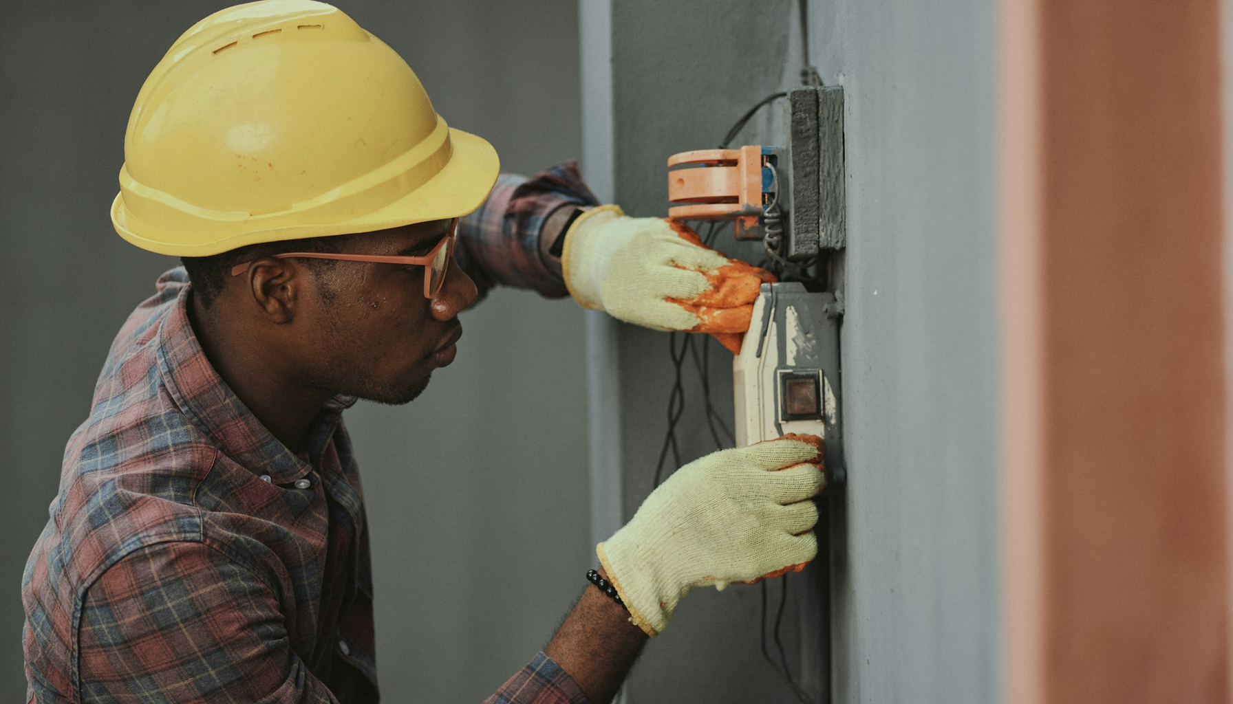 An electrician in a yellow hard hat and work gloves installing wiring on a wall