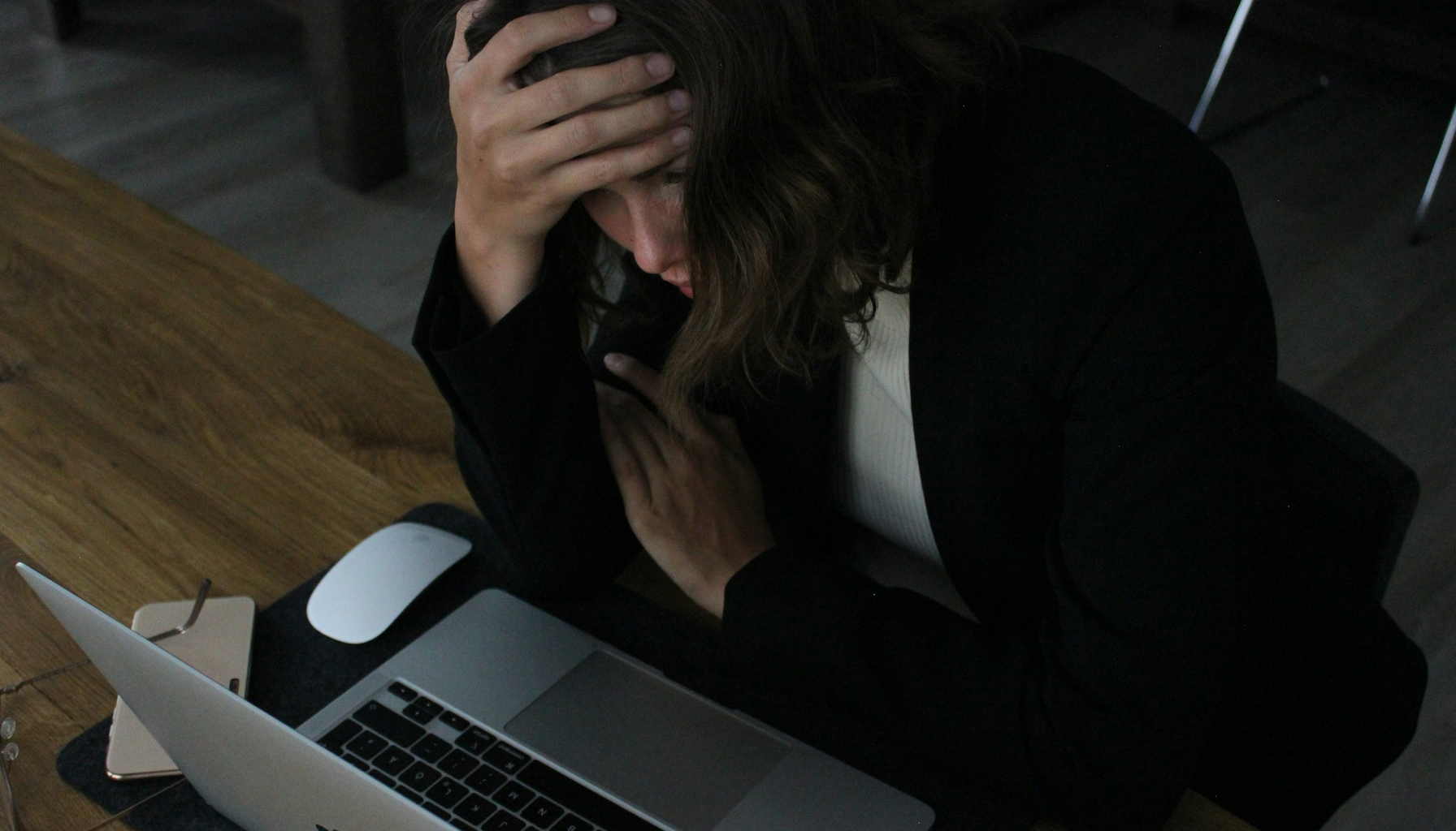 A woman with her head in her hand, frustrated, looking at a laptop on a wooden desk